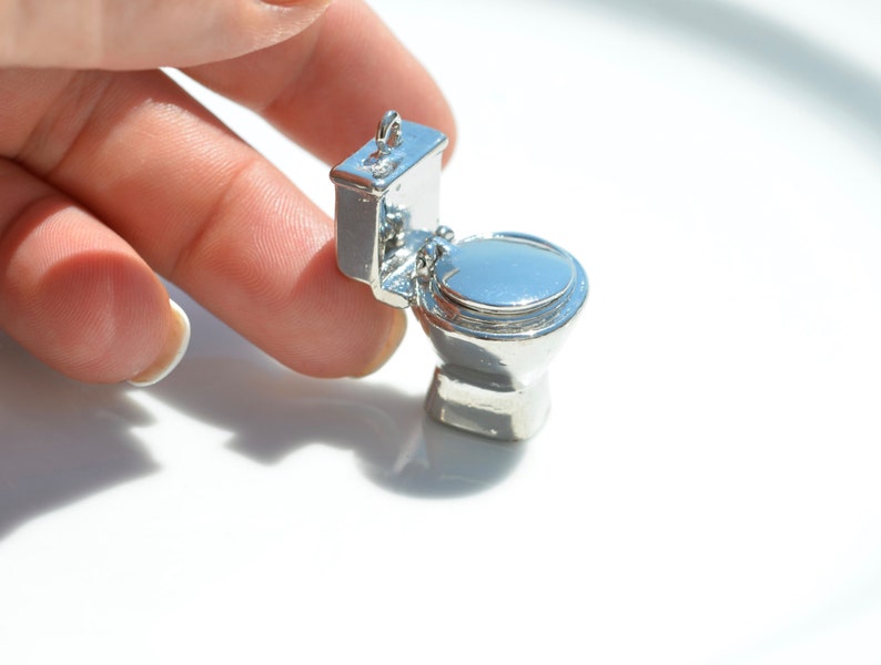 May include: A silver-colored miniature toilet charm. The toilet is open and the lid is up. The charm is small enough to fit on a finger.