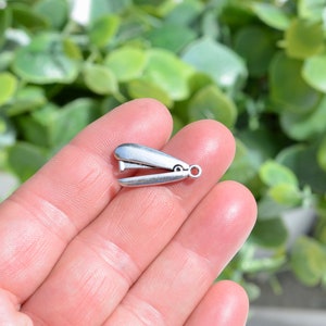 May include: A small, silver-colored miniature stapler charm. The charm is held in a hand, showcasing its detailed design and small size. The stapler has a loop for attaching to jewelry or other crafts.