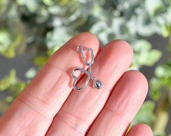 May include: Silver stethoscope charm with a heart-shaped loop. The charm is on a hand.