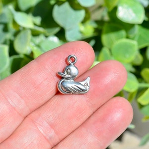 May include: Silver charm in the shape of a rubber duck. The duck is facing to the right and has a small hole at the top for attaching to a chain or bracelet.
