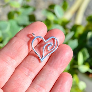May include: Silver heart-shaped pendant with a swirl design inside. The pendant is held in a hand.