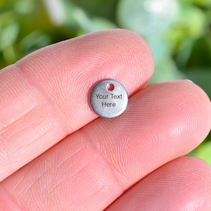 May include: A small, round, silver-colored charm with a hole at the top. The charm has the text "Your Text Here" printed on its surface. The charm is held between two fingers against a blurred green background.