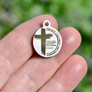 May include: A circular silver-toned metal pendant featuring a dark brown brushstroke cross design. The text "PRAY ON IT, PRAY OVER IT, PRAY THROUGH IT" circles the edge, with "In all your ways acknowledge Him, And He shall direct your paths. Proverbs 3:6" printed in the centre.