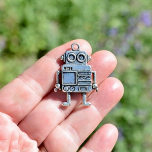 May include: Silver robot charm with a simple design. The robot has two round eyes, a rectangular body with a wavy line, and two small arms and legs.