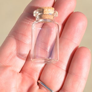 May include: A small, clear glass bottle with a cork stopper. The bottle is cylindrical with a rounded base and a narrow neck. The cork is light brown. The bottle is held in a person's hand.