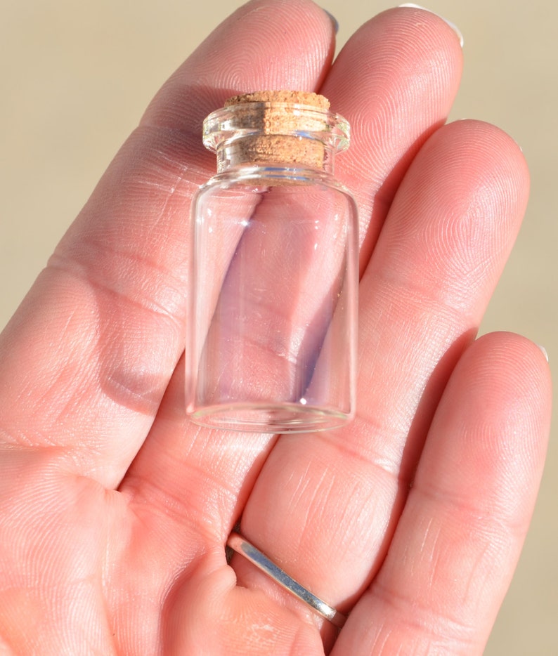 May include: A small, clear glass bottle with a cork stopper. The bottle is cylindrical with a rounded base and a narrow neck. The cork is light brown. The bottle is held in a person's hand.