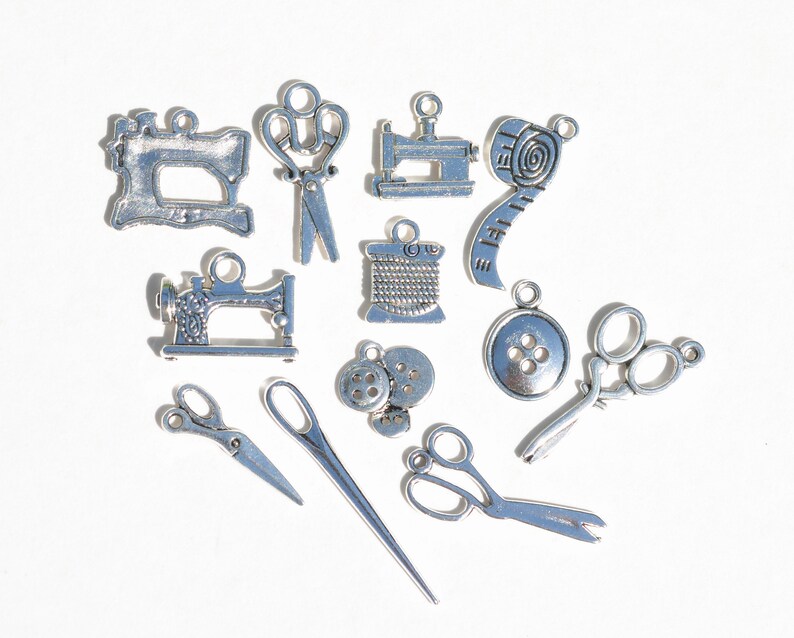 May include: A collection of silver-toned metal charms with sewing themes. The charms include a sewing machine, scissors, buttons, a spool of thread, and a measuring tape.