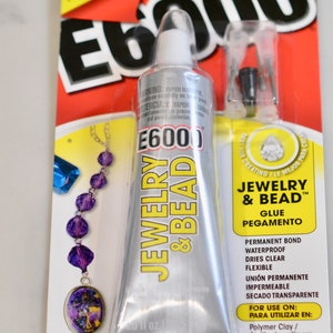 Op de afbeelding: Een zilveren tube E6000 Jewelry & Bead lijm met een geel etiket. Het etiket vermeldt "Permanente Binding, Waterdicht, Droogt helder, Flexibel" en "Voor gebruik op: Polymer Clay, Metaal, Hout, Glas".