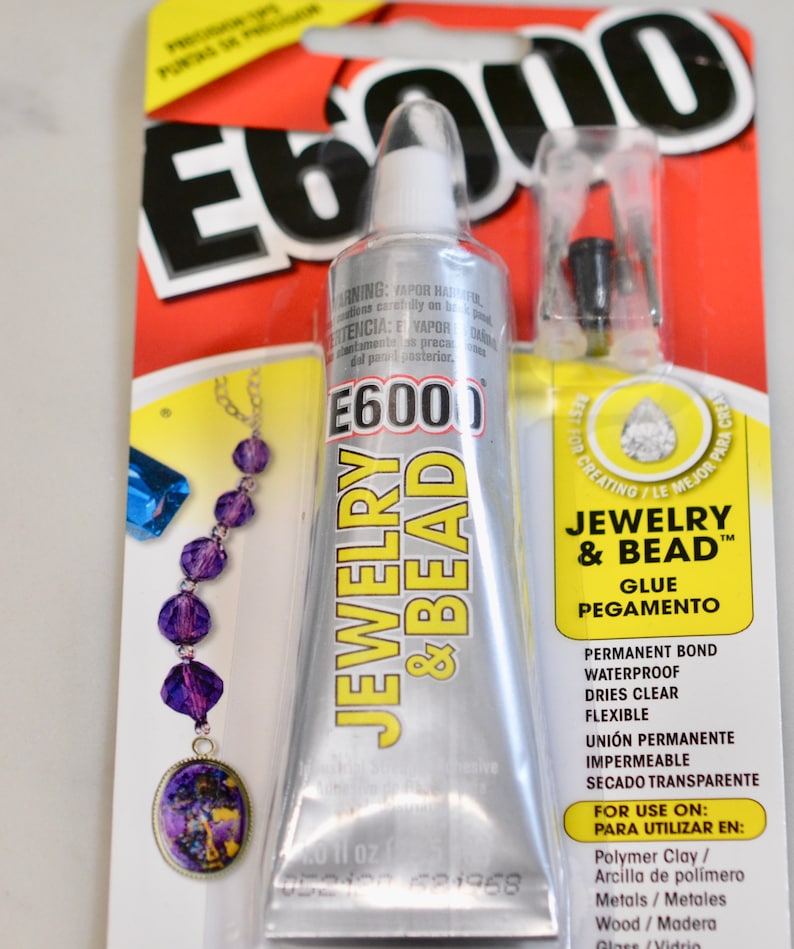Op de afbeelding: Een zilveren tube E6000 Jewelry & Bead lijm met een geel etiket. Het etiket vermeldt "Permanente Binding, Waterdicht, Droogt helder, Flexibel" en "Voor gebruik op: Polymer Clay, Metaal, Hout, Glas".