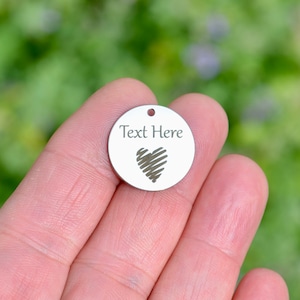 May include: A round silver charm with a heart design and the text "Text Here" engraved on it.