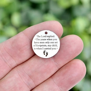 May include: A circular silver-toned metal charm featuring a small hole at the top. The surface is engraved with black text that reads: The Lord replied: The years when you have seen only one set of footprints, my child, is when I carried you.