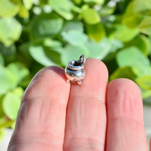 May include: A small, silver-colored cauldron charm with three tiny feet and a loop at the top. The charm is held in a person's hand, with a blurred green background.