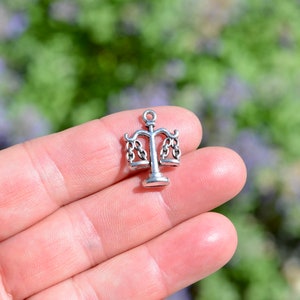May include: A silver charm in the shape of a scale of justice. The charm is small and detailed, with two hanging scales and a base.
