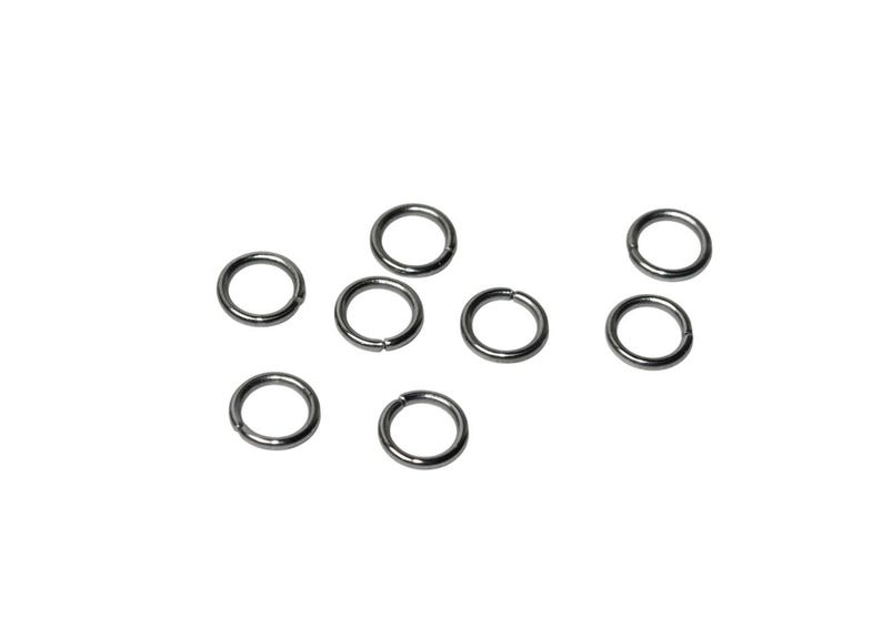 May include: A collection of seven small, circular metal jump rings with a dark gunmetal finish. Each ring features a single, clean cut along its circumference, allowing the ring to be opened and closed for connecting jewelry components.