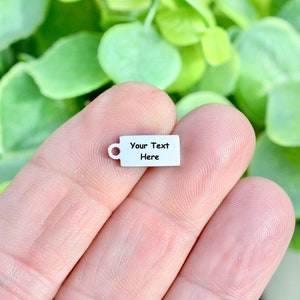 May include: A small, silver-colored charm in the shape of a coffee mug. The charm has a rectangular body with a handle on the side and the text "Your Text Here" printed on the front. The charm is held in a person's hand.