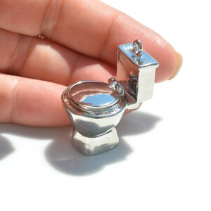 May include: A silver charm in the shape of a toilet. The toilet is in a closed position with the lid down. The charm is small enough to fit in the palm of a hand.