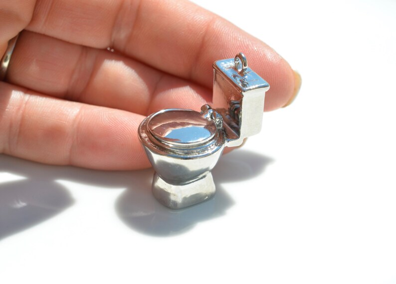 May include: A silver charm in the shape of a toilet. The toilet is in a closed position with the lid down. The charm is small enough to fit in the palm of a hand.