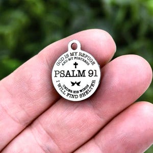 May include: A silver-colored round pendant with the text "GOD IS MY REFUGE AND MY FORTRESS PSALM 91 I WILL FIND UNDER HIS WINGS SHELTER" encircling a cross and a bow tie design. The pendant has a small loop at the top.