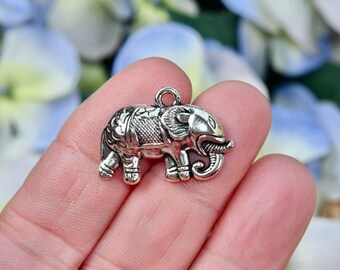 Op de afbeelding: Een kleine, zilverkleurige olifantenbedel met gedetailleerde gravures. De bedel heeft een lus aan de bovenkant om aan sieraden of andere knutselwerken te bevestigen. De slurf van de olifant is gekruld en zijn oren zijn prominent.
