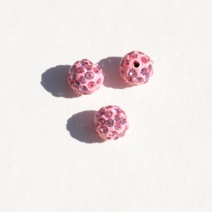 May include: Three pink round beads with a sparkly finish. Each bead is covered in small pink rhinestones.