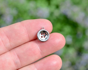 May include: A small, shiny silver button charm with four holes and a loop at the top. The button is round and has a polished finish. The background is a soft, blurred green and white.