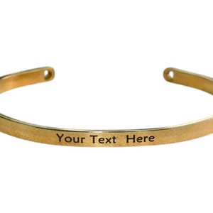May include: A gold-colored cuff bracelet with the engraved text "Your Text Here". The bracelet has a simple, open design with rounded edges and small holes at each end. This piece of jewelry is suitable for personalization.