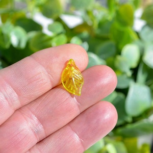 May include: A yellow glass leaf charm with a detailed vein pattern. The charm is translucent and has a hole at the top for hanging.