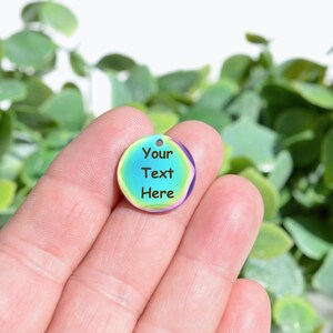May include: A circular, iridescent charm with a hole at the top, held in a hand. The charm has a blue and green centre with the text "Your Text Here" in black. The edge of the charm is purple.