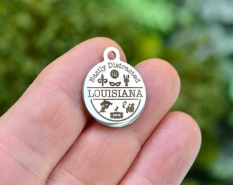 May include: A circular silver-coloured metal tag featuring the engraved text "Easily Distracted" and "LOUISIANA". The design includes icons of a fleur-de-lis, a mask, a crawfish, a swamp tree, a streetcar, a rabbit, and a saxophone player.