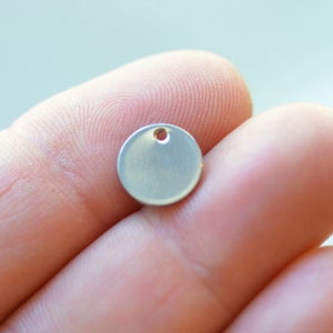 May include: A small, circular, silver-colored metal charm with a smooth, matte finish. The disc features a single, centered hole near the top edge, designed for attachment to jewelry or other items. The surface is plain and unadorned.