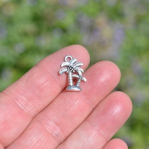 May include: Silver charm featuring a pair of palm trees with detailed leaves and trunks. The charm is designed to be used for jewelry making or crafting projects.