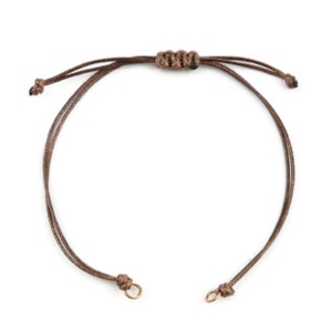 May include: A brown adjustable bracelet with a sliding knot closure. The bracelet features two thin cords that form a loop, with small gold-colored metal rings at the ends. The bracelet is designed to be adjustable to fit various wrist sizes.