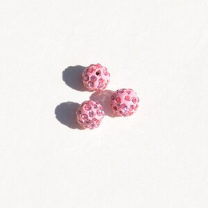 May include: Three pink round beads with a sparkly finish. Each bead is covered in small pink rhinestones.