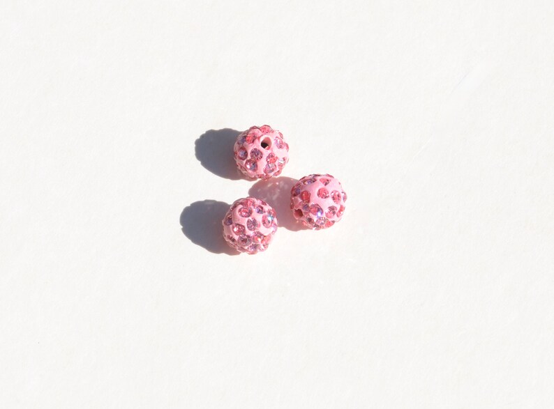 May include: Three pink round beads with a sparkly finish. Each bead is covered in small pink rhinestones.