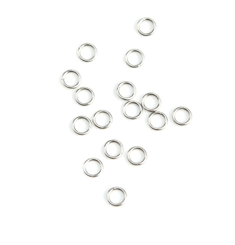 May include: A scattering of silver jump rings on a white background. The rings are small and circular, and they are arranged in a random pattern.