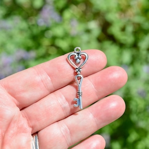 May include: A silver key charm featuring a heart-shaped design at the top and a keyhole. The key is held in a hand against a blurred green and purple backdrop.