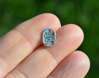 May include: A small, silver-colored metal charm with the words "MADE WITH LOVE" embossed on it. The charm is oval-shaped with a small hole at the top for stringing. The background is a blurred green.