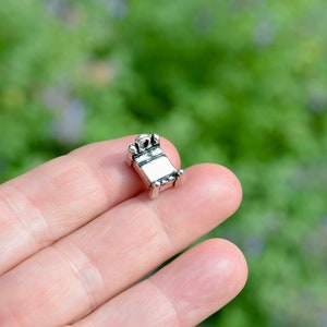 May include: Silver charm shaped like a small bed with a detailed design. The charm is being held in a hand.