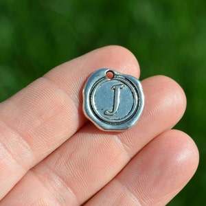 May include: Silver charm with a raised letter J in a circle. The charm has a textured surface and a hole for attaching to a chain or bracelet.