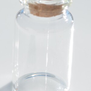 May include: A small, clear glass bottle with a cork stopper. The bottle is cylindrical with a rounded base and a narrow neck. The cork is a light brown color and fits snugly into the bottle's opening. The bottle is empty.