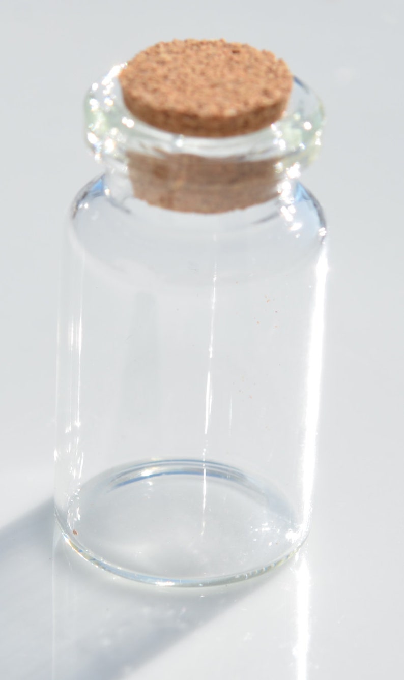 May include: A small, clear glass bottle with a cork stopper. The bottle is cylindrical with a rounded base and a narrow neck. The cork is a light brown color and fits snugly into the bottle's opening. The bottle is empty.