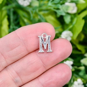 May include: Silver charm in the shape of the letter M. The charm has a loop at the top for attaching to a bracelet or necklace.