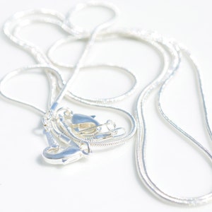 Puede incluir: Un collar de cadena de serpiente de plata con dos cierres. La cadena est&aacute; enrollada y colocada sobre una superficie blanca.