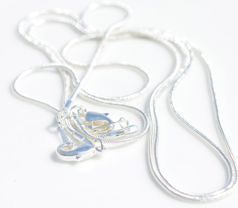 Puede incluir: Un collar de cadena de serpiente de plata con dos cierres. La cadena est&aacute; enrollada y colocada sobre una superficie blanca.