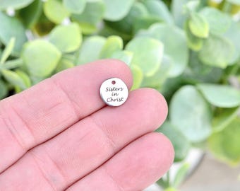 May include: A small, circular silver charm bearing the inscription "Sisters in Christ". The charm, with a small hole at the top, is held in a hand. The background features blurred green foliage.