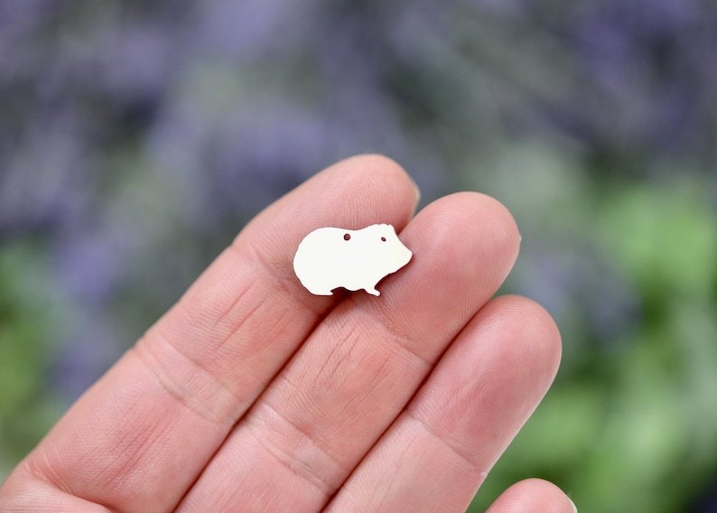 May include: A white metal charm shaped like a guinea pig. The charm has two small holes for attaching to a necklace or bracelet.