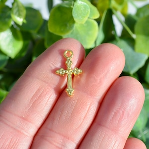 May include: A small gold-colored cross charm with a detailed design. The cross is held in a hand against a green background.