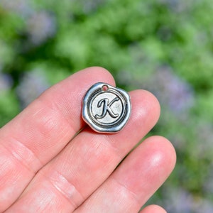 May include: Silver charm with a wax seal design featuring the letter K.