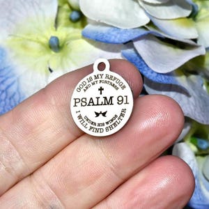 May include: A round, silver-colored charm with the text "GOD IS MY REFUGE AND MY FORTRESS PSALM 91 I WILL FIND SHELTER UNDER HIS WINGS". The charm has a small loop at the top for attaching to a necklace or bracelet.