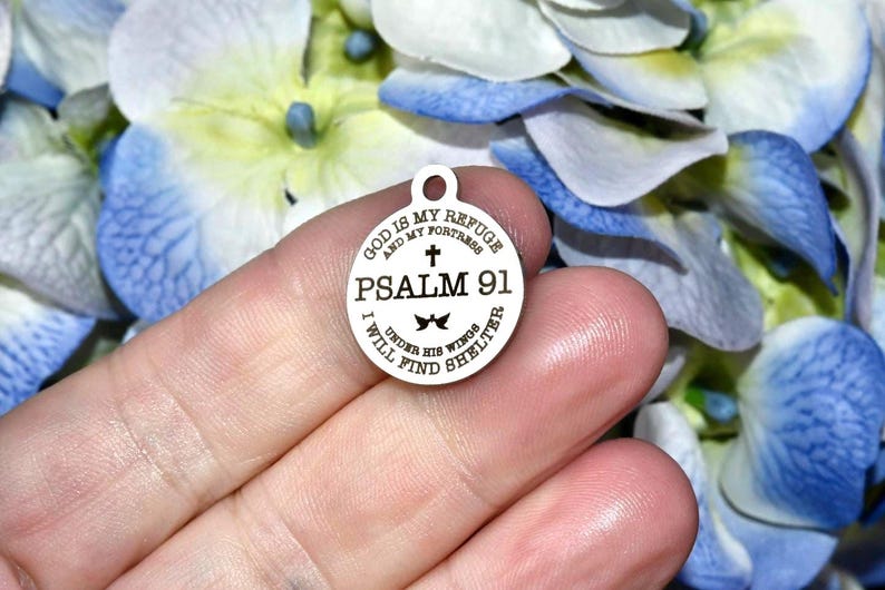 May include: A round, silver-colored charm with the text "GOD IS MY REFUGE AND MY FORTRESS PSALM 91 I WILL FIND SHELTER UNDER HIS WINGS". The charm has a small loop at the top for attaching to a necklace or bracelet.